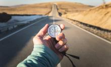 Photo of an extended hand holding a compass in front of a highway that stretches into the distance.