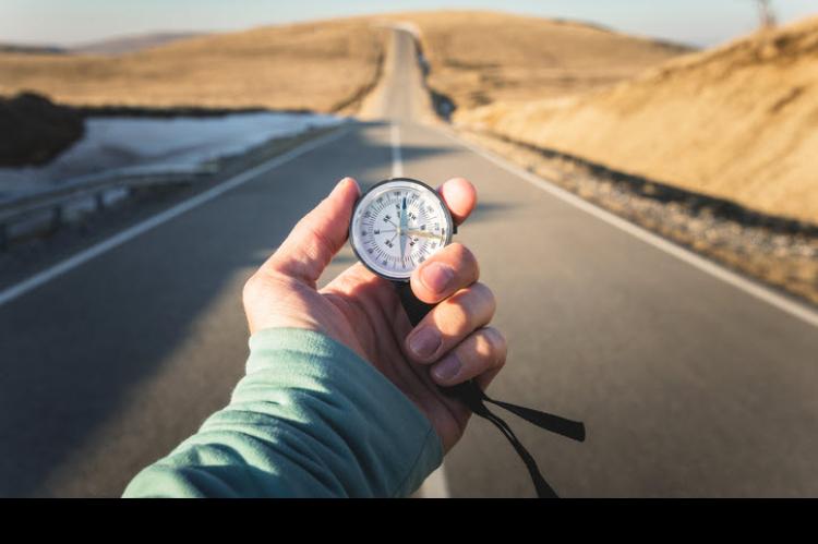 Photo of an extended hand holding a compass in front of a highway that stretches into the distance.