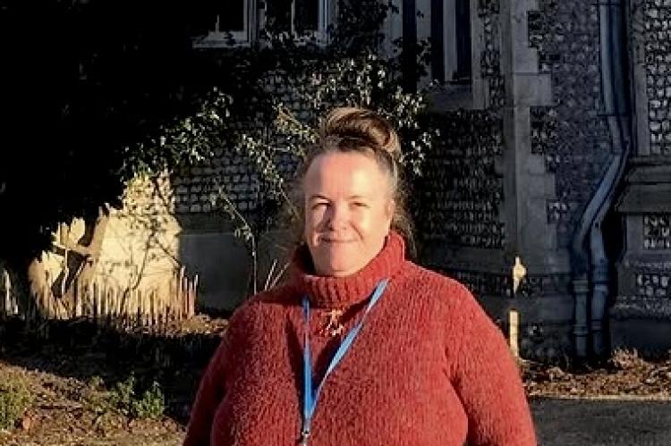 Anne Welsh in April 2024, wearing a red rollneck sweater and smiling, with an old building in the background