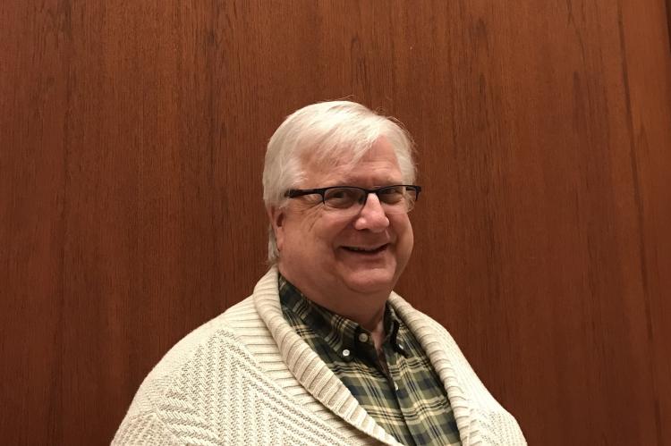 Robert Maxwell, standing in front of a wood-panelled wall. He is wearing a cream cardigan and green and cream-checked shirt, and dark, half-rimmed glasses, and is smiling into the camera. He has short, white hair.