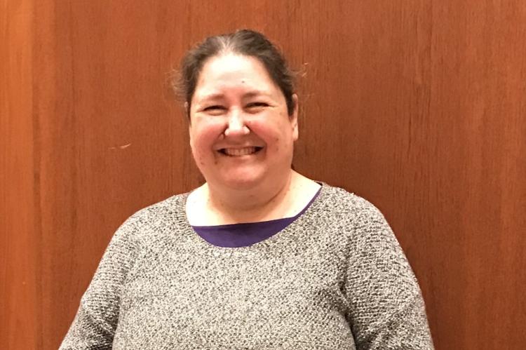 Charlotte Christensen, standing against a wall with wood panelling. She us wearing a grey marl jumper and is smiling at the camera. She has long, dark hair.