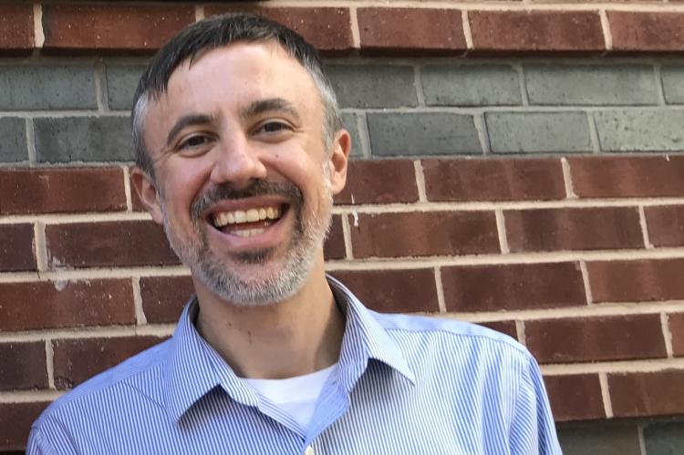 Chris Holden, standing in front of a brick wall, smiling into the camera and wearing a pale blue and white striped shirt