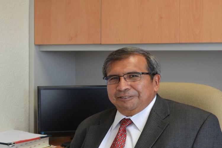 Filiberto Felipe Martinez Arellano, a man with salt and pepper hair and glasses, is sitting at a modern desk, wearing a grey suit, white shirt and red tie, and is smiling at the camera.