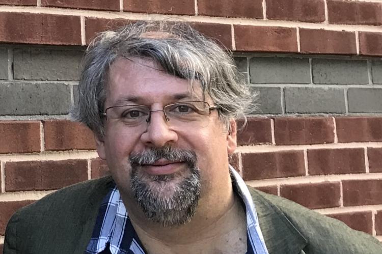 Scabolcs Dancs, smiling, wearing a grey suit jacket and blue checked shirt, standing against a brick wall