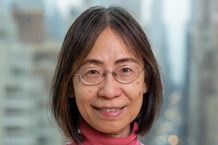 Charlene Chou - a woman with dark hair, wearing glasses and a pink rollneck jumper, smiling at the camera and standing before a blurred-out cityscape (New York)