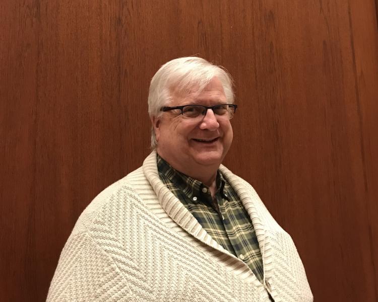 Robert Maxwell, standing in front of a wood-panelled wall. He is wearing a cream cardigan and green and cream-checked shirt, and dark, half-rimmed glasses, and is smiling into the camera. He has short, white hair. 