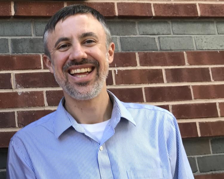 Chris Holden, standing in front of a brick wall, smiling into the camera and wearing a pale blue and white striped shirt