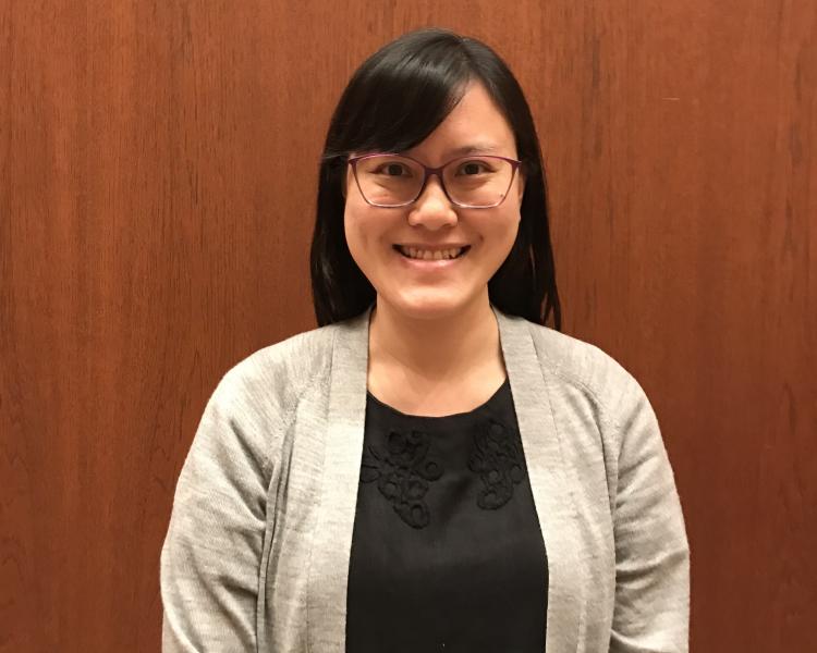 Elisa, smiling at the camera, wearing red-framed glasses, a black dress and grey cardigan, and standing in front of a wood-panelled door.