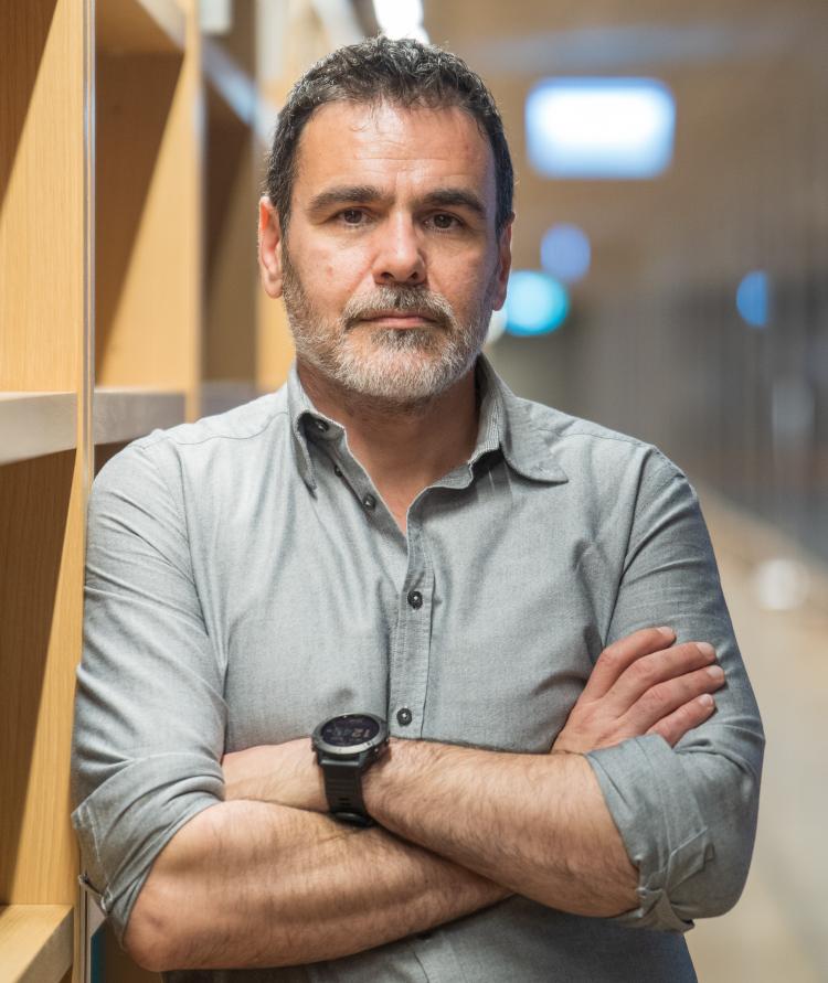 Michalis Gerolimos (National Library of Greece). A man with dark hair and eyes and a neatly-trimmed grey beard looks straight into the camera. In the background some library shelves and some lights have been blurred out of focus.
