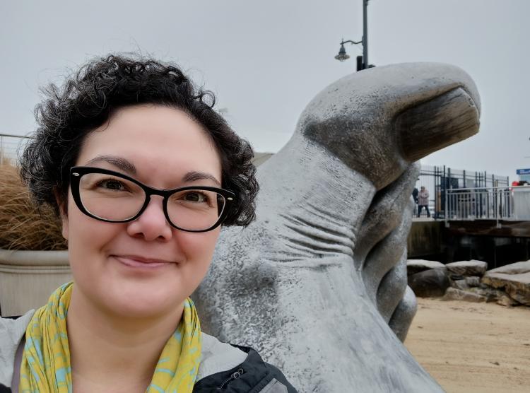 Trina Soderquist, a woman with curly black hair and dark eyes wearing black-framed glasses, smiling into the camera, in front of a modern sculpture of a foot.