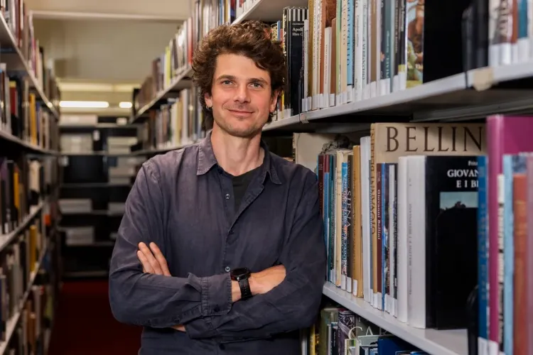 Jack Ennis Butler, a man with dark wavy hair, wearing a blue shirt, staring directly into the camera and smiling, leans against a shelf in a stack of books.