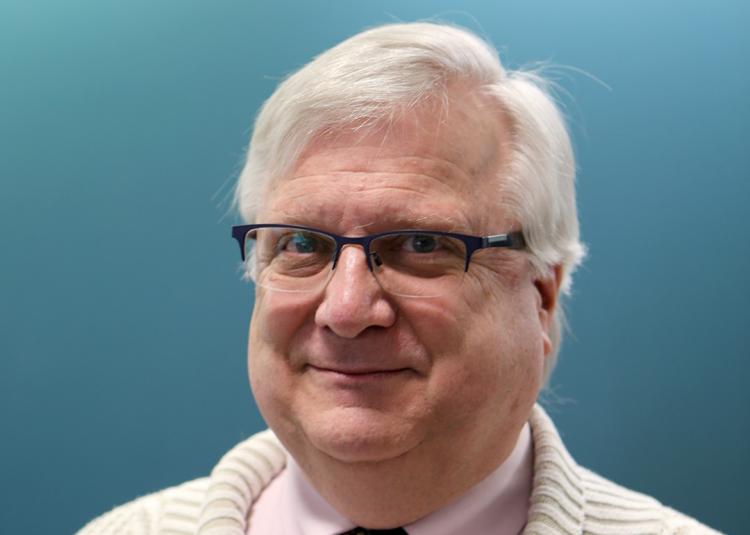 Landscape version of Robert L. Maxwell's headshot - a man with silver hair and horn-rimmed glasses smiling into the camera. He is wearing a cream shirt and cardigan and is photograped against a blue background.