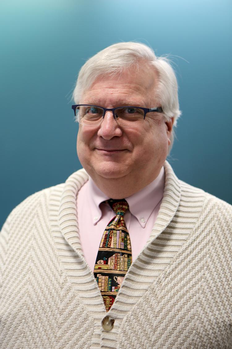 Robert L. Maxwell, smiling into the camera, wearing a cream shirt and cardigan and a tie featuring shelves of books. He has silver hair and horn-rimmed spectacles.