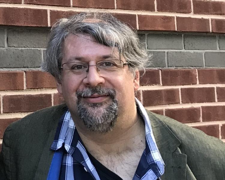 Scabolcs Dancs, smiling, wearing a grey suit jacket and blue checked shirt, standing against a brick wall