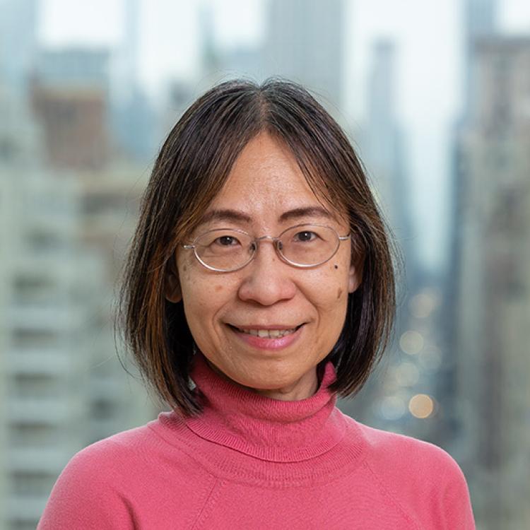 Charlene Chou - a woman with dark hair, wearing glasses and a pink rollneck jumper, smiling at the camera and standing before a blurred-out cityscape (New York)