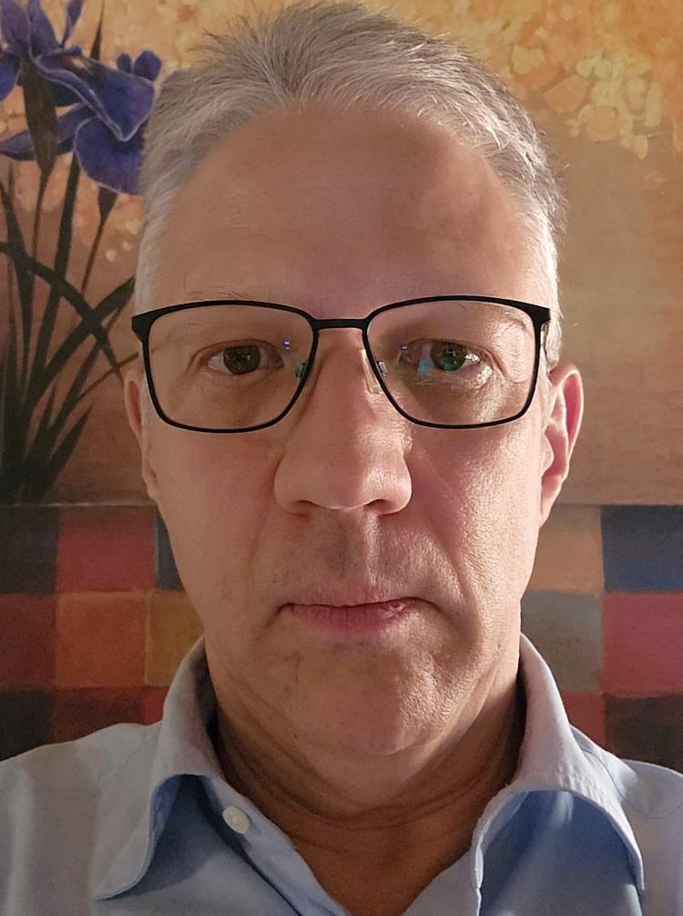 Thomas Brenndorfer, a man with silver hair and black-rimmed glasses, smiles at the camera. In the background are colourful tiles and a painting of a bearded iris.
