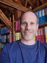 Martin Luepold, a man with a short beard, smiling into the camera. He is standing in front of a run of wooden shelves with colour-co-ordinated ring binders. 
