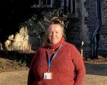 Anne Welsh in April 2024, wearing a red rollneck sweater and smiling, with an old building in the background
