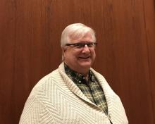 Robert Maxwell, standing in front of a wood-panelled wall. He is wearing a cream cardigan and green and cream-checked shirt, and dark, half-rimmed glasses, and is smiling into the camera. He has short, white hair. 