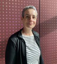 Bridget Jennings, a woman with close-cropped grey hair, wearing a grey striped top and a black jacket, stands in front of a mauve background with light mauve stars, smiles into the camera