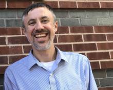Chris Holden, standing in front of a brick wall, smiling into the camera and wearing a pale blue and white striped shirt