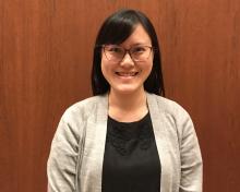 Elisa, smiling at the camera, wearing red-framed glasses, a black dress and grey cardigan, and standing in front of a wood-panelled door.