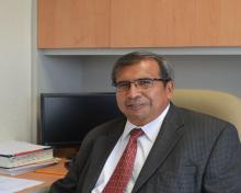 Filiberto Felipe Martinez Arellano, a man with salt and pepper hair and glasses, is sitting at a modern desk, wearing a grey suit, white shirt and red tie, and is smiling at the camera.