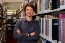 Jack Ennis Butler, a man with dark wavy hair, wearing a blue shirt, staring directly into the camera and smiling, leans against a shelf in a stack of books.