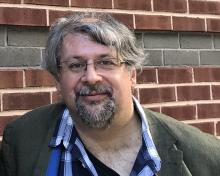 Scabolcs Dancs, smiling, wearing a grey suit jacket and blue checked shirt, standing against a brick wall