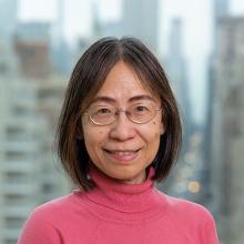 Charlene Chou - a woman with dark hair, wearing glasses and a pink rollneck jumper, smiling at the camera and standing before a blurred-out cityscape (New York)